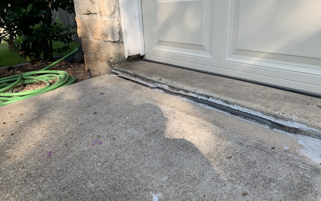 A photo showing a driveway with a noticeable gap between the concrete and the garage entrance, likely due to sinking or settling. The gap has been filled with a white sealant, which extends along the length of the joint.